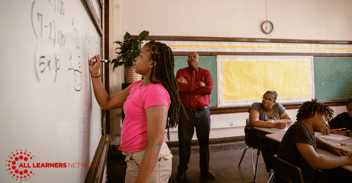 Female student writing on a whiteboard as a teacher and other students look on